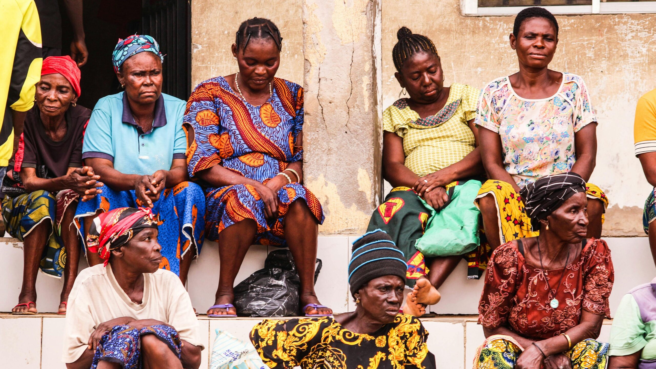 A group of African women in colorful traditional dresses sit on stairs, reflecting everyday urban life.